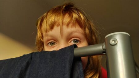 beautiful little girl is hiding back bunk bed which dark blue velvet fabric hangs child has gray eyes blond hair upper half face is visible hide seek wp
